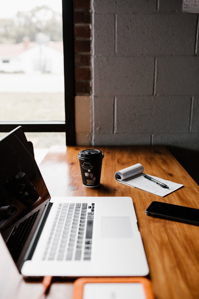 Modern office desk setup with laptop, coffee cup, and notepad by window.